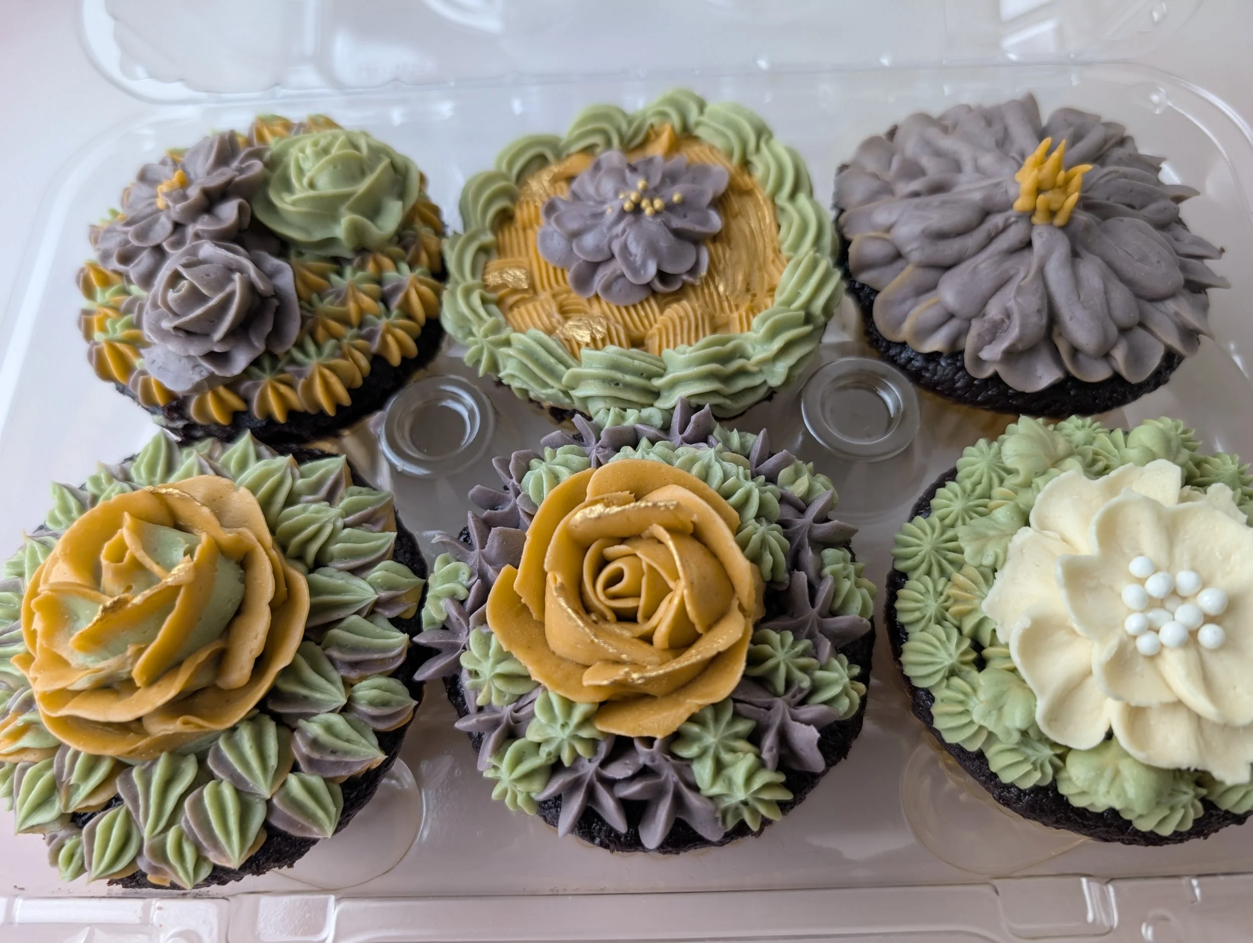 Set of decorated cupcakes with various yellow, green, purple, and cream-colored frosting designs arranged in a clear plastic container.