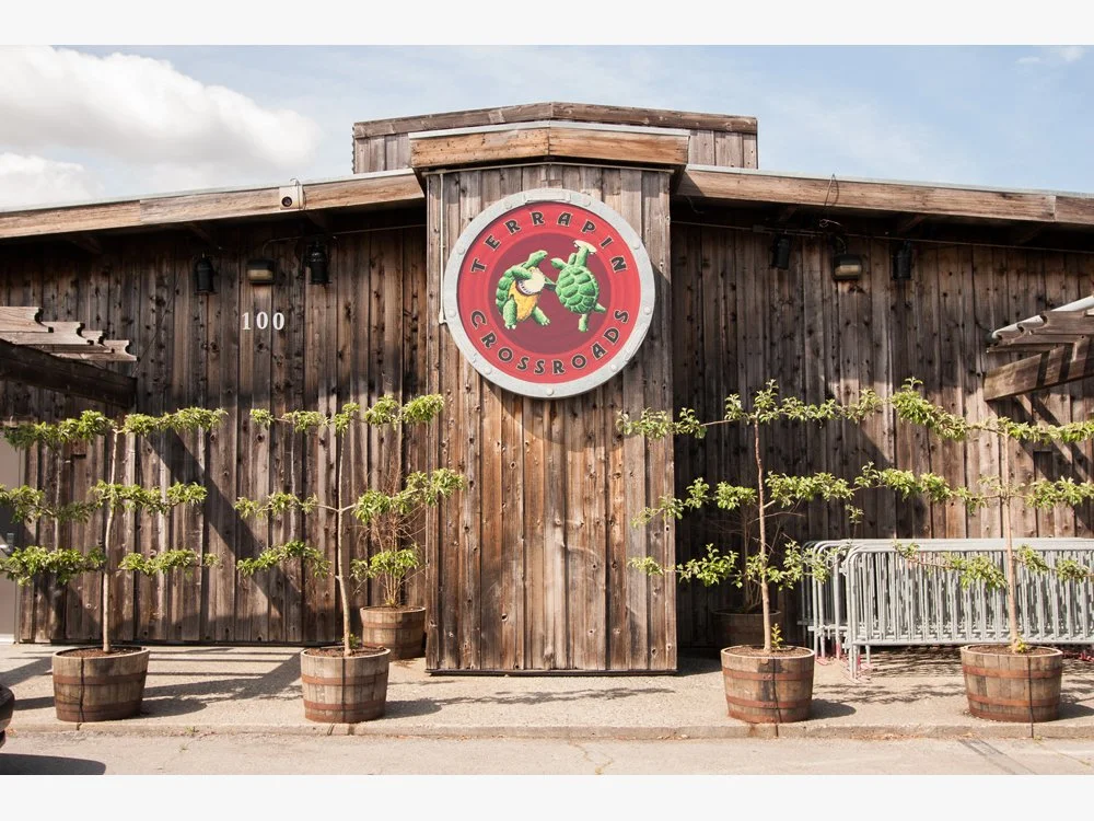 Wooden building with a circular sign that reads 'Terrapin Crossroads' and features a drawing of a turtle. There are five potted trees in front of the building and some metal barricades on the right.