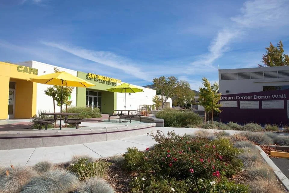 A colorful outdoor area with a yellow and green building labeled 'SAY DREAM CENTER,' yellow umbrellas, picnic tables, and a purple wall with white text labeled 'Dream Center Donor Wall.'