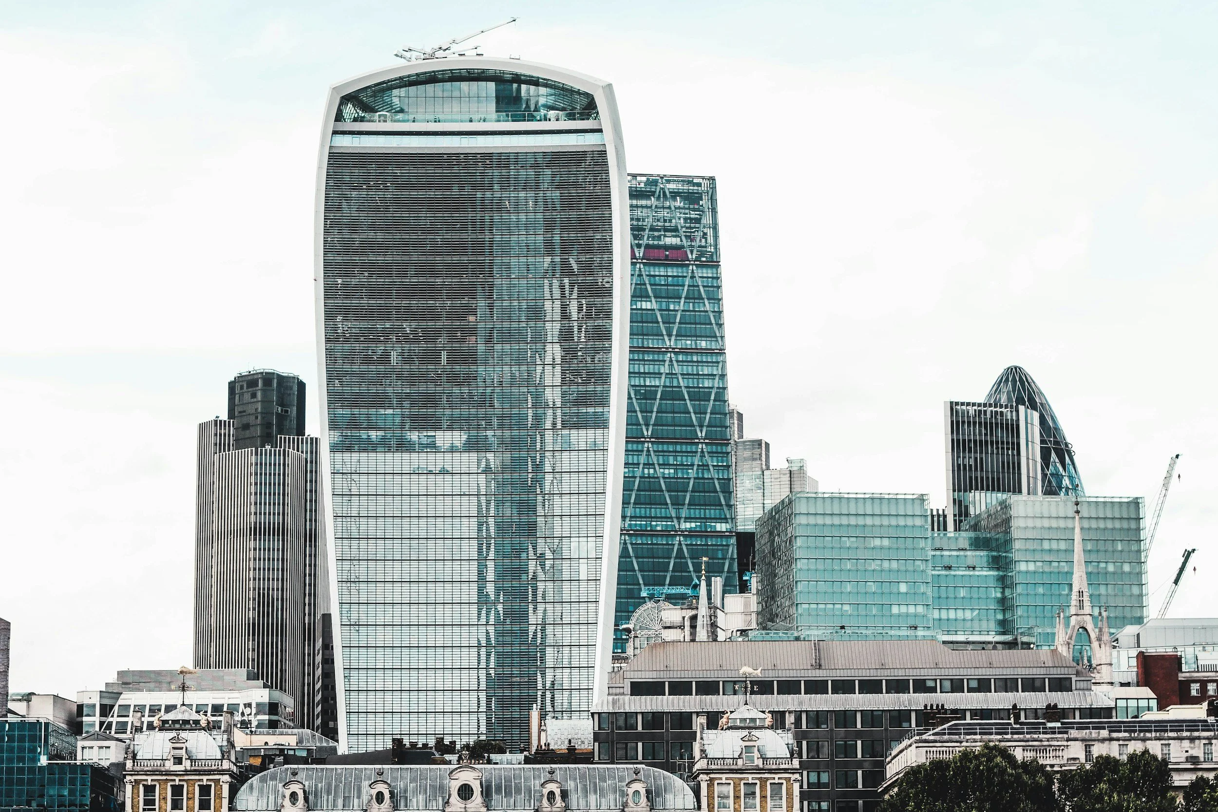 City skyline featuring modern skyscrapers, including the prominent Walkie Talkie building with a curved glass facade, and other high-rise office buildings against a cloudy sky.