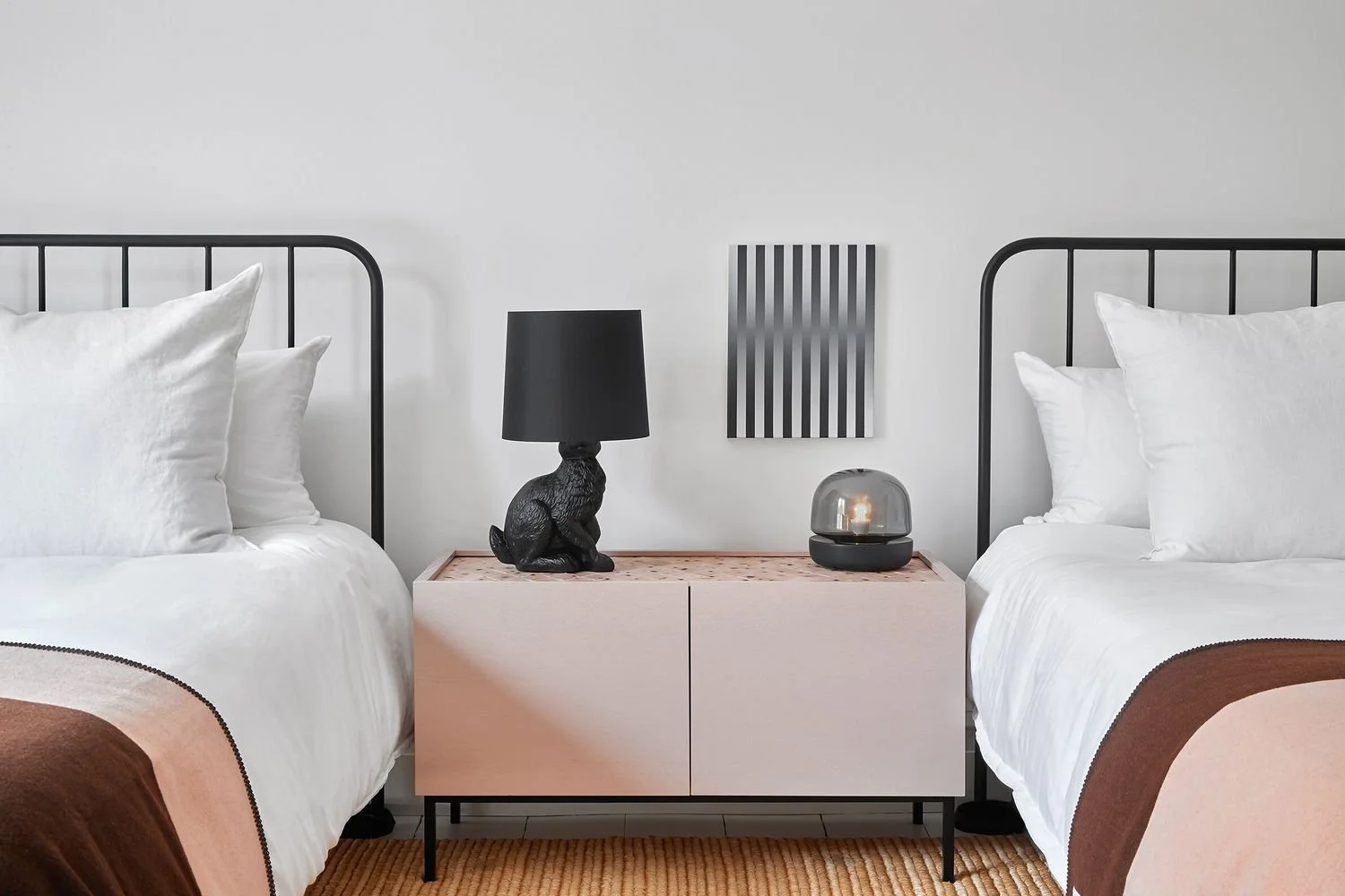 A bedroom with two beds having white linens and black metal headboards, a pink bedside table with a black rabbit-shaped lamp with a black lampshade, a black and silver decorative object, and a black and white striped artwork on a white wall.