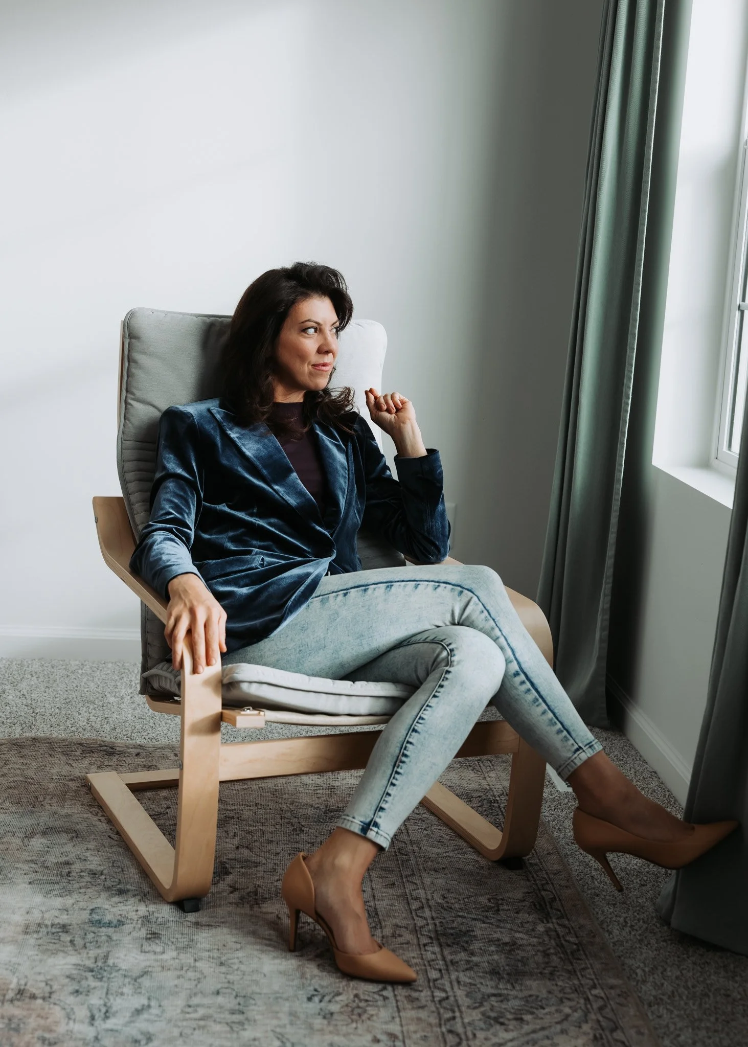 A woman sitting in a modern chair, looking out a window with gray curtains, wearing a dark blue velvet blazer, light jeans, and nude high heels.