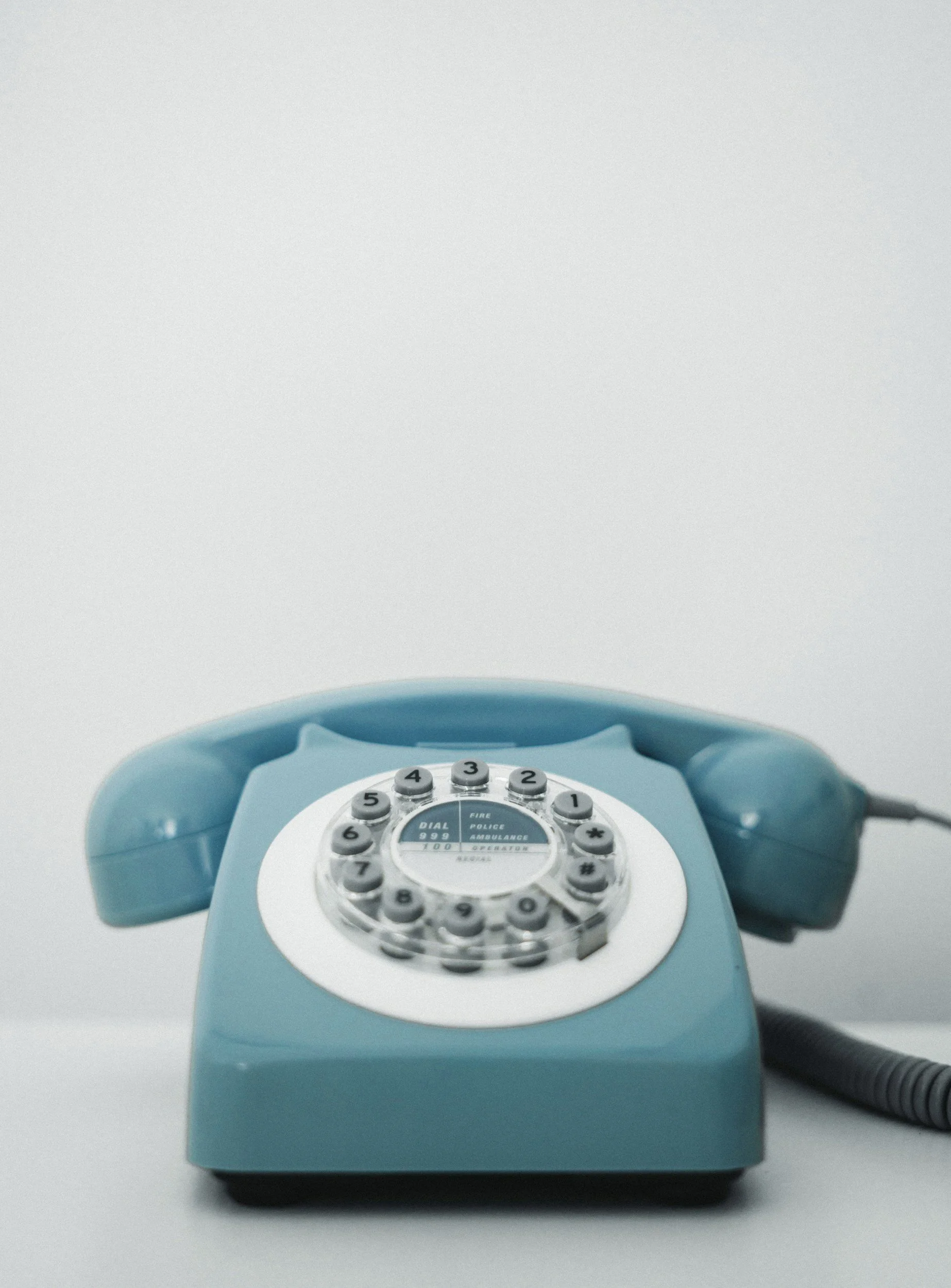 A vintage rotary telephone in light blue color resting on a white surface with a plain white background.