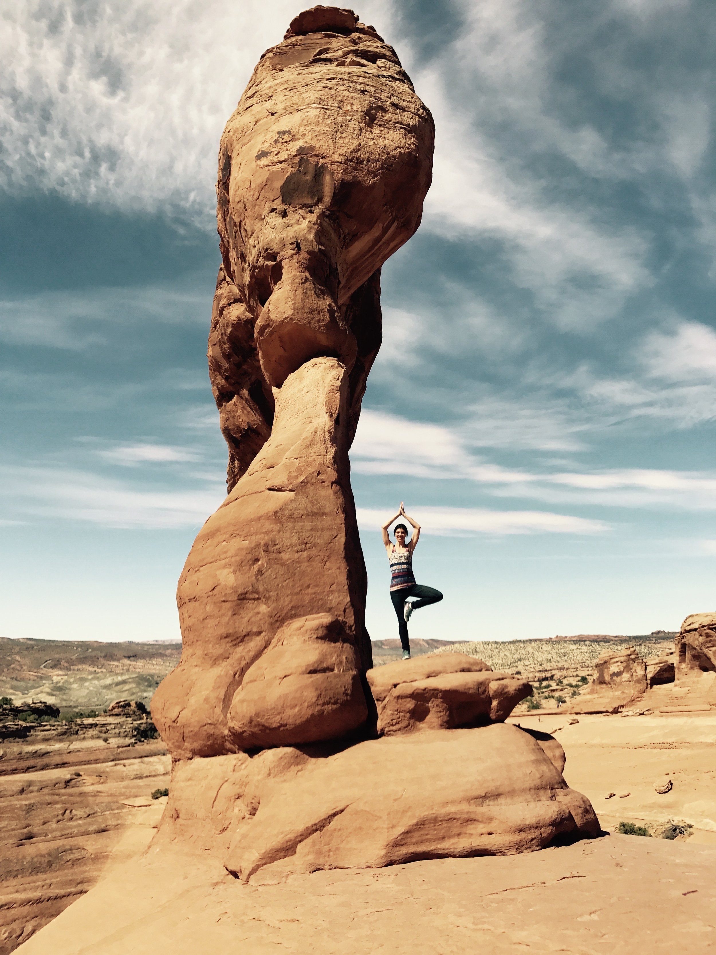 A person practicing yoga in a tree pose next to a tall rock formation in a desert landscape.