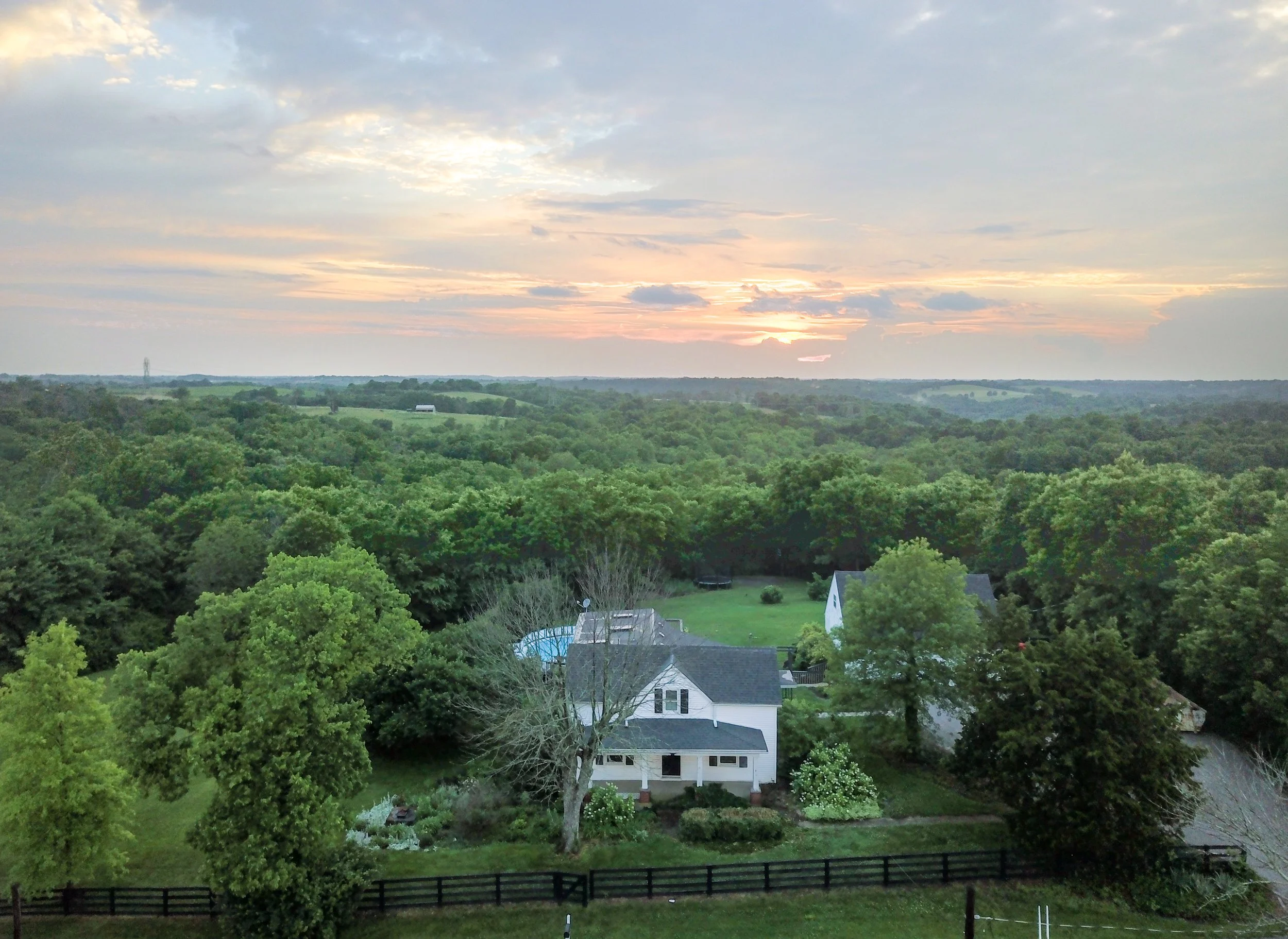 A scenic view of a rural neighborhood during sunset with houses, green trees, and rolling hills under a partly cloudy sky.