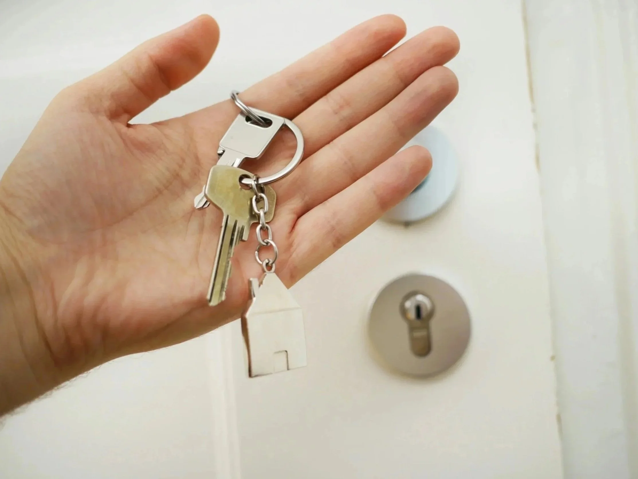 Close-up of a person's hand holding a keychain with several keys, positioned near a white wall with electrical outlets and a silver lock.