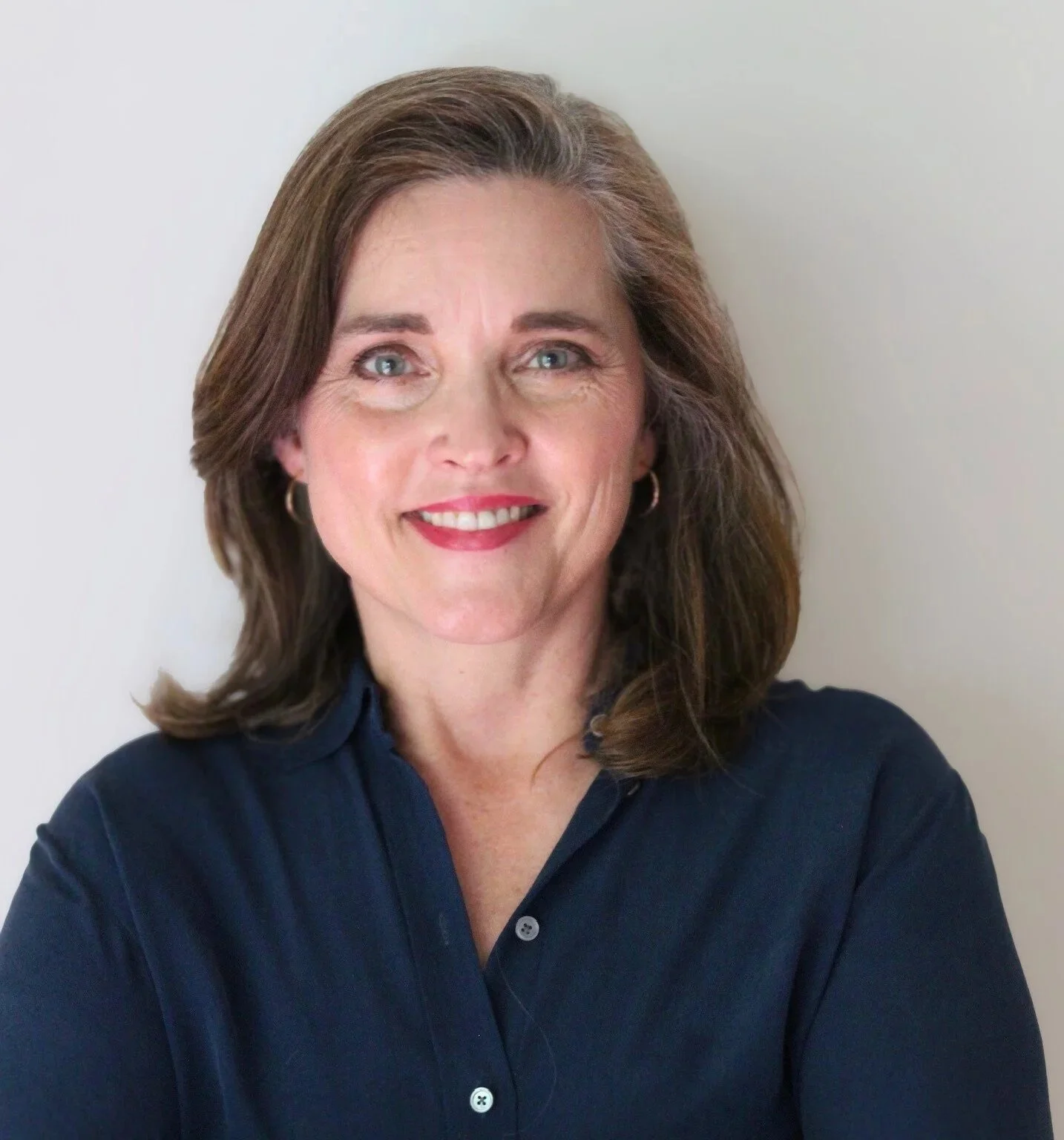 A woman with shoulder-length brown hair, blue eyes, wearing a dark blue blouse, smiling, standing against a plain white wall.