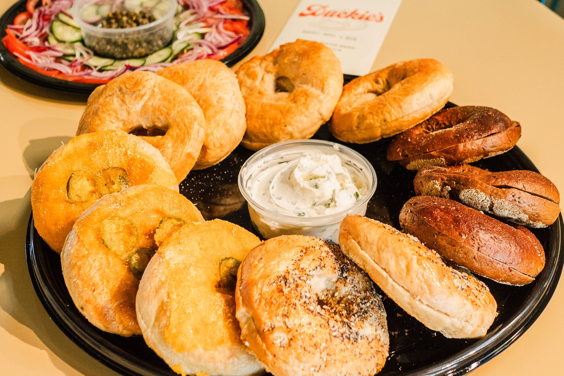 A platter of assorted bagels, including sesame, plain, and multigrain, served with cream cheese, alongside a side salad of cucumber, tomato, and onion slices with a small container of dipping sauce in the background.