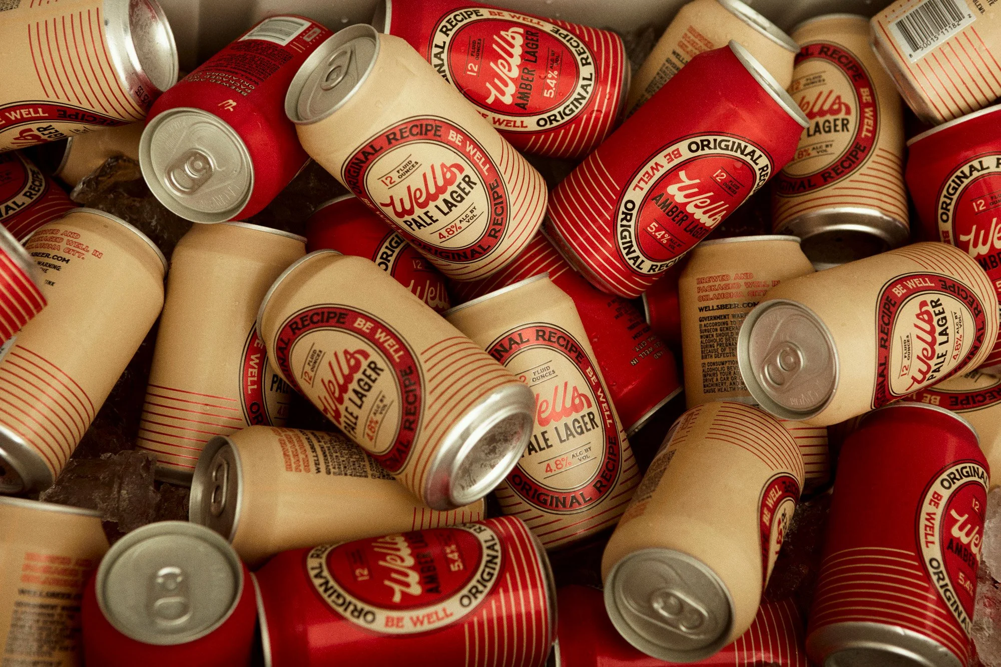 A pile of well used cans of Wells Pale Lager beer with red and beige labels, some with pull tabs, and ice underneath.