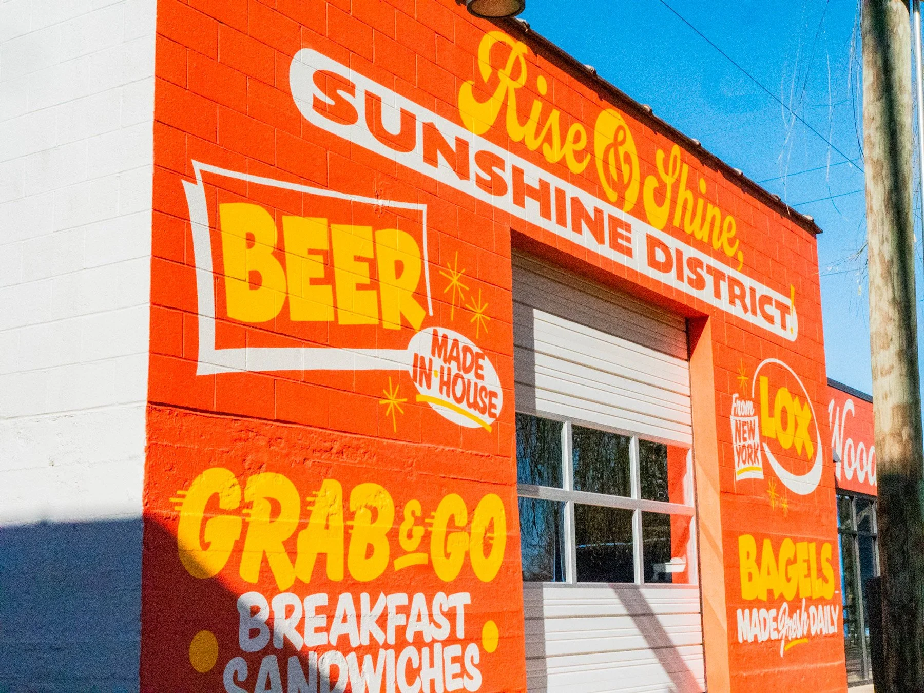 Brightly colored mural of a bakery storefront with signs advertising beer, bagels, breakfast sandwiches, and other baked goods. The background is painted orange with yellow, white, and red lettering.