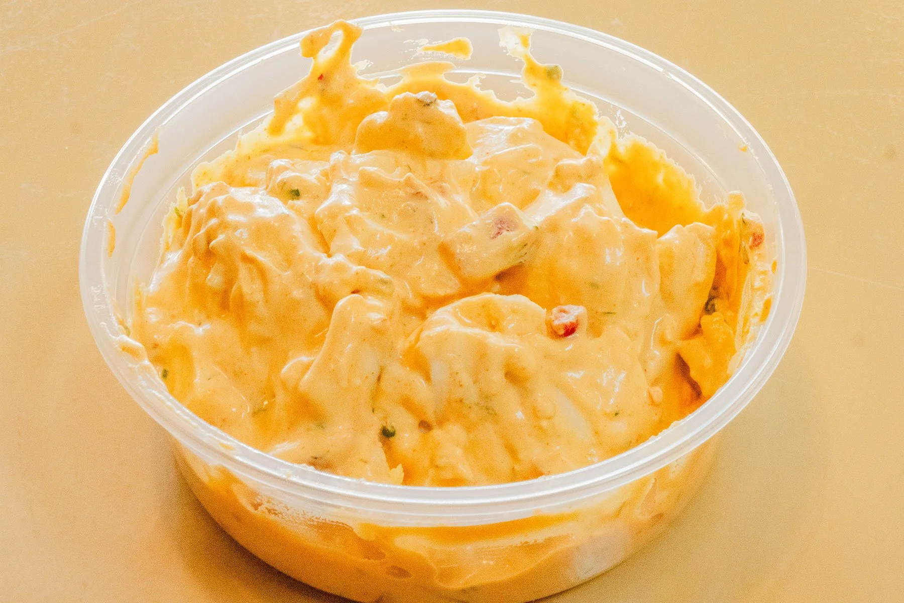 A clear plastic container filled with creamy potato salad with chunks of potatoes, mayonnaise, and small bits of vegetables on a beige surface.