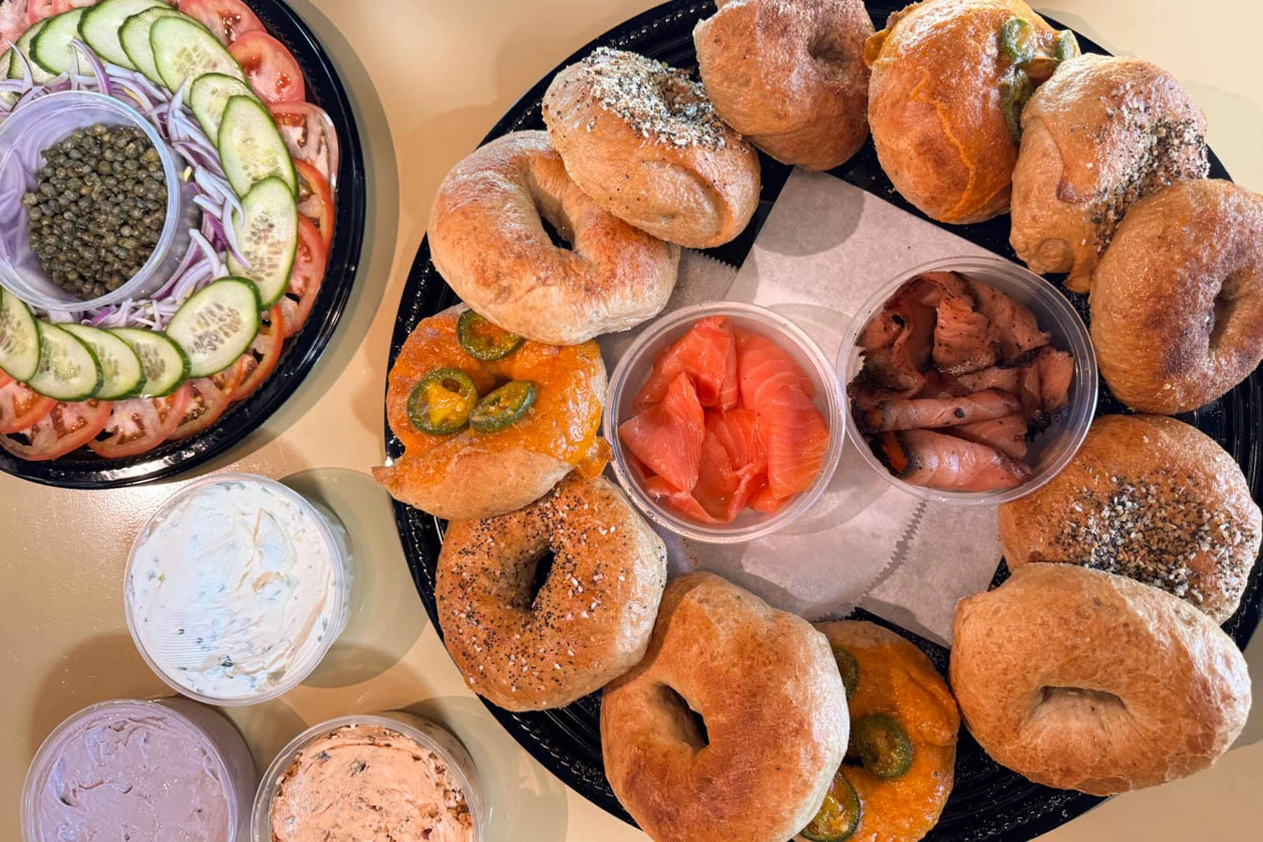 A plate of assorted baked bread rolls, some topped with sesame or poppy seeds, and containers of smoked salmon, pickled vegetables, and various whipped spreads or dips, with a side salad of sliced cucumbers, tomatoes, red onions, and a bowl of green peppercorns.