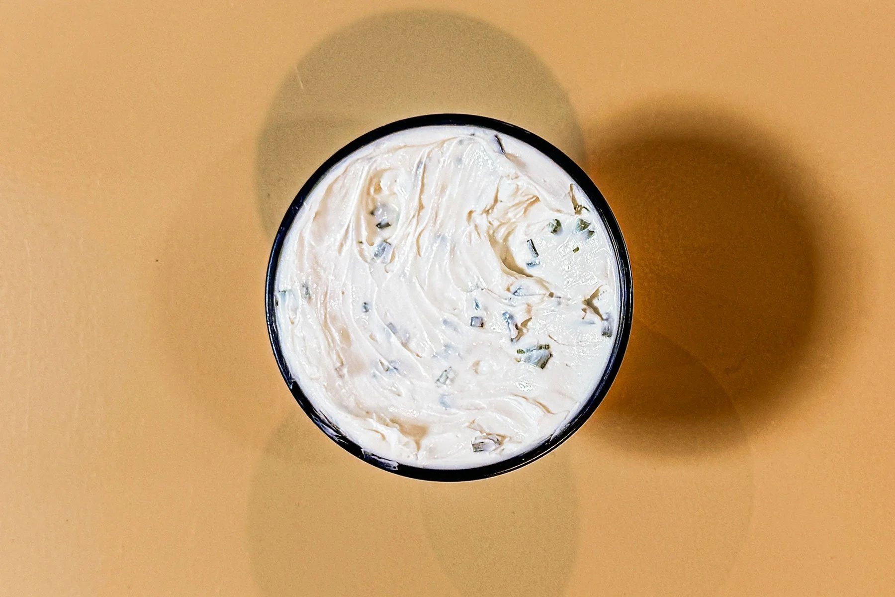 A close-up top view of an opened container of creamy, white substance with small green flecks, on a plain beige background.