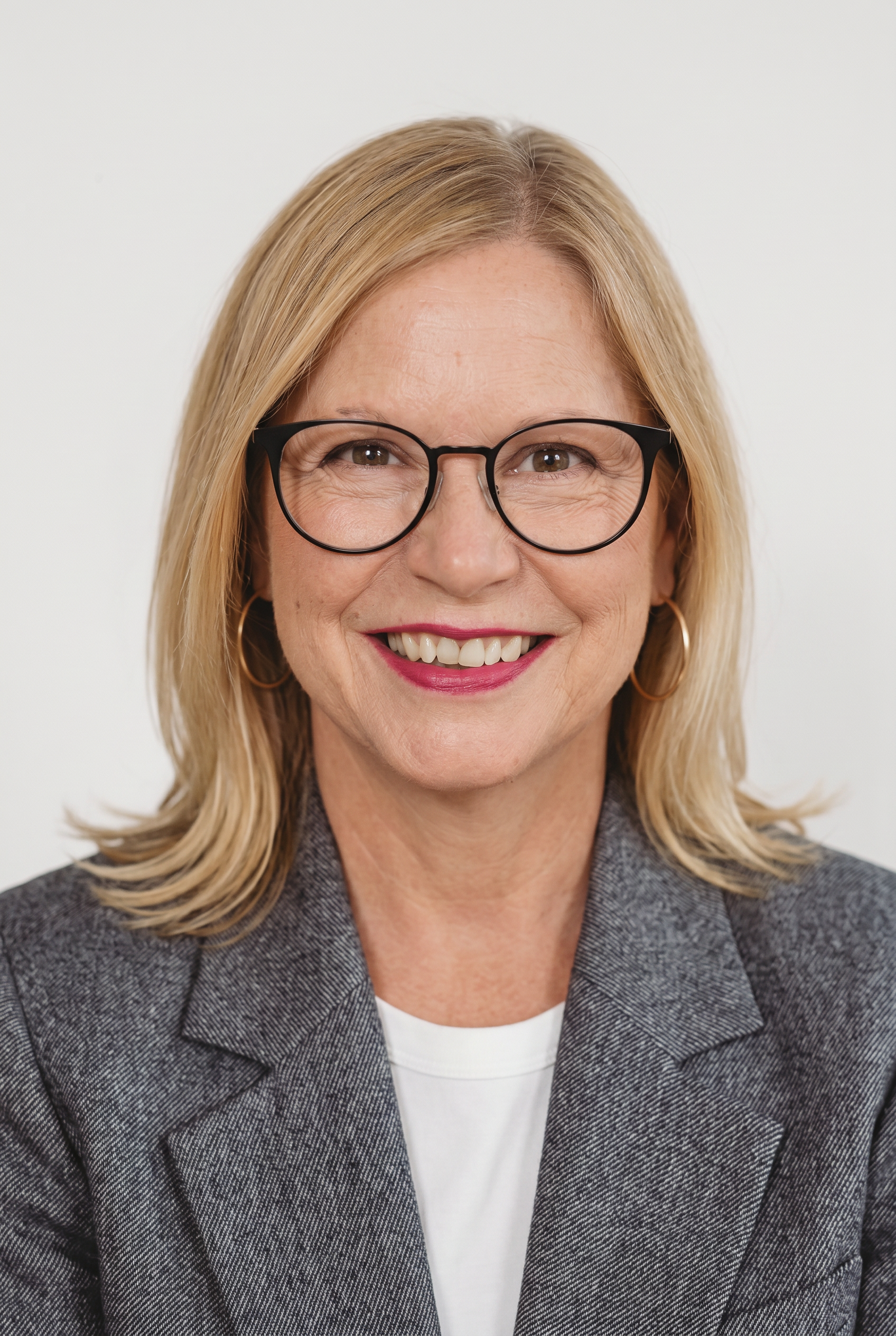 Porträt einer lächelnden Frau mit blonden Haaren, Brille, grauem Hosenmantel und weißen Top vor weißem Hintergrund.