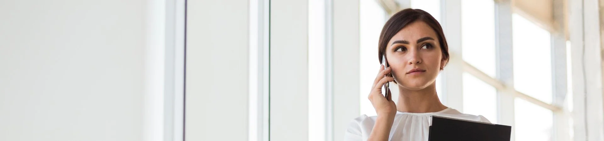 A woman in business attire talking on her phone while holding a tablet, standing near large windows with natural light.