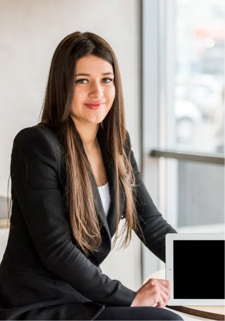 A young woman with long brown hair, wearing a black blazer, sitting at a desk near a large window with a white tablet in her hand.