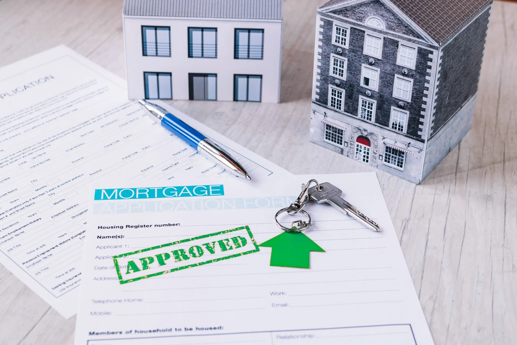 Mortgage application form with a green approved stamp, keys, and small house models on a wooden surface.