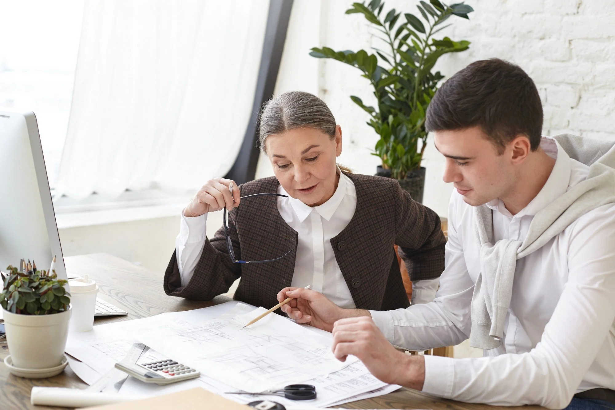 Two people discussing architectural plans at a desk, with a computer, calculator, and potted plants nearby.