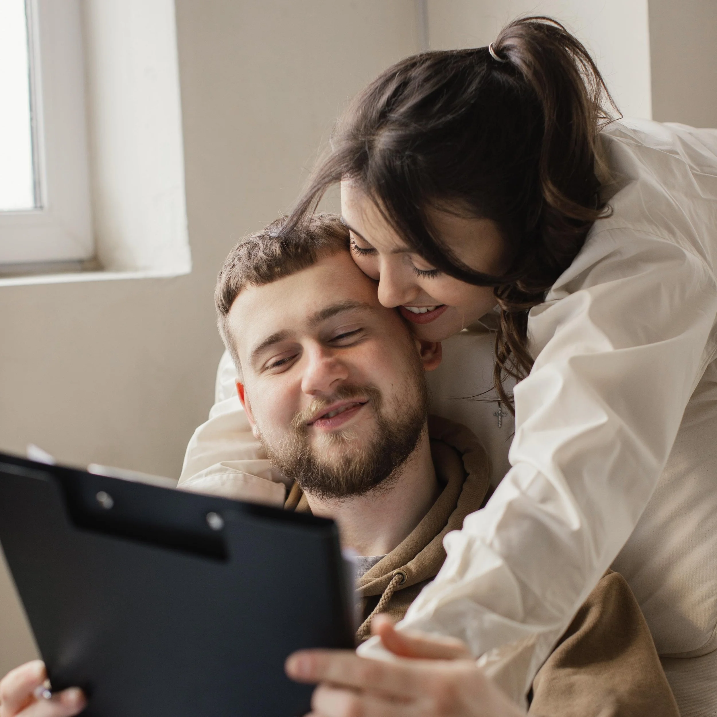 A woman leans over a man sitting in bed, both smiling and looking at a tablet device together.