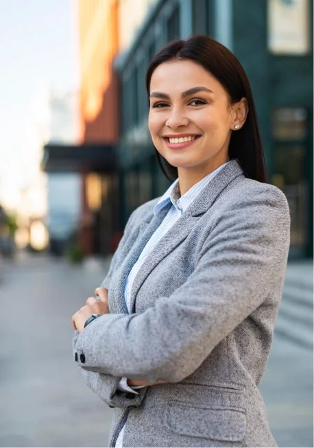 A smiling woman in a light gray business suit standing outdoors in an urban setting with modern glass buildings.