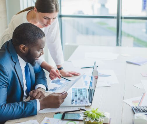 Two professionals, a man and a woman, reviewing documents and discussing work in an office setting with natural light.