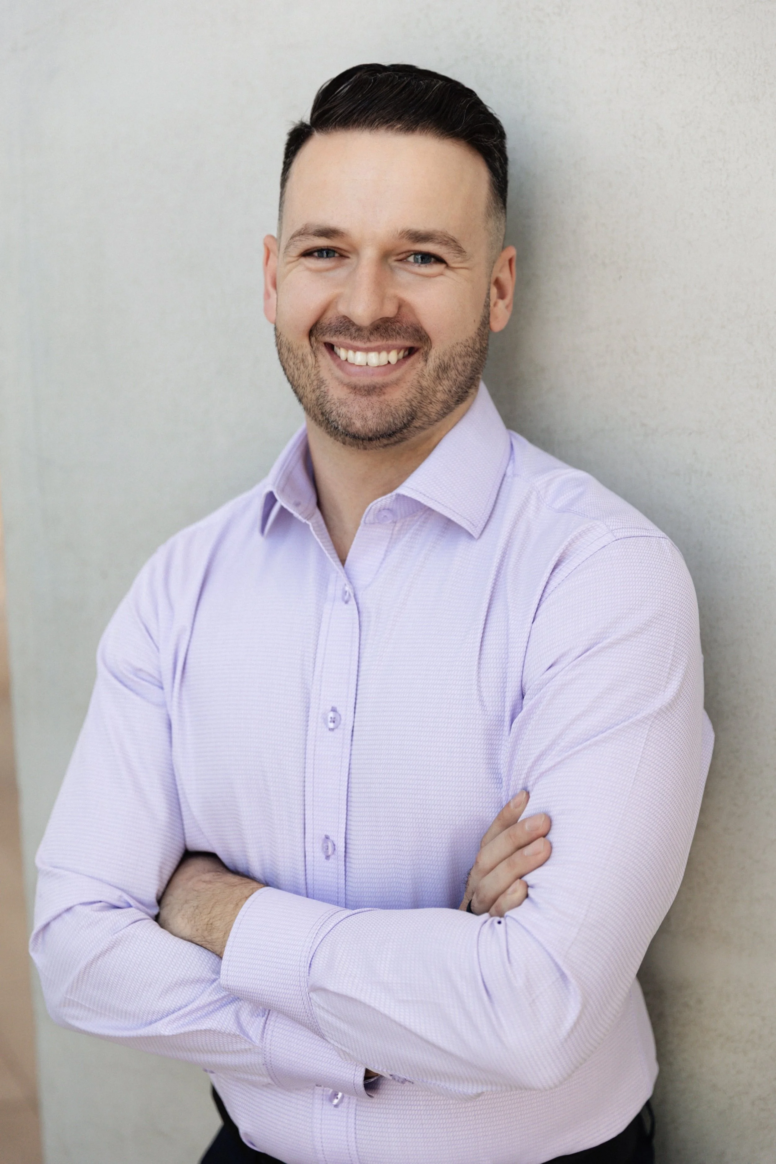 A smiling man in a light purple button-up shirt crossing his arms, standing against a neutral background.