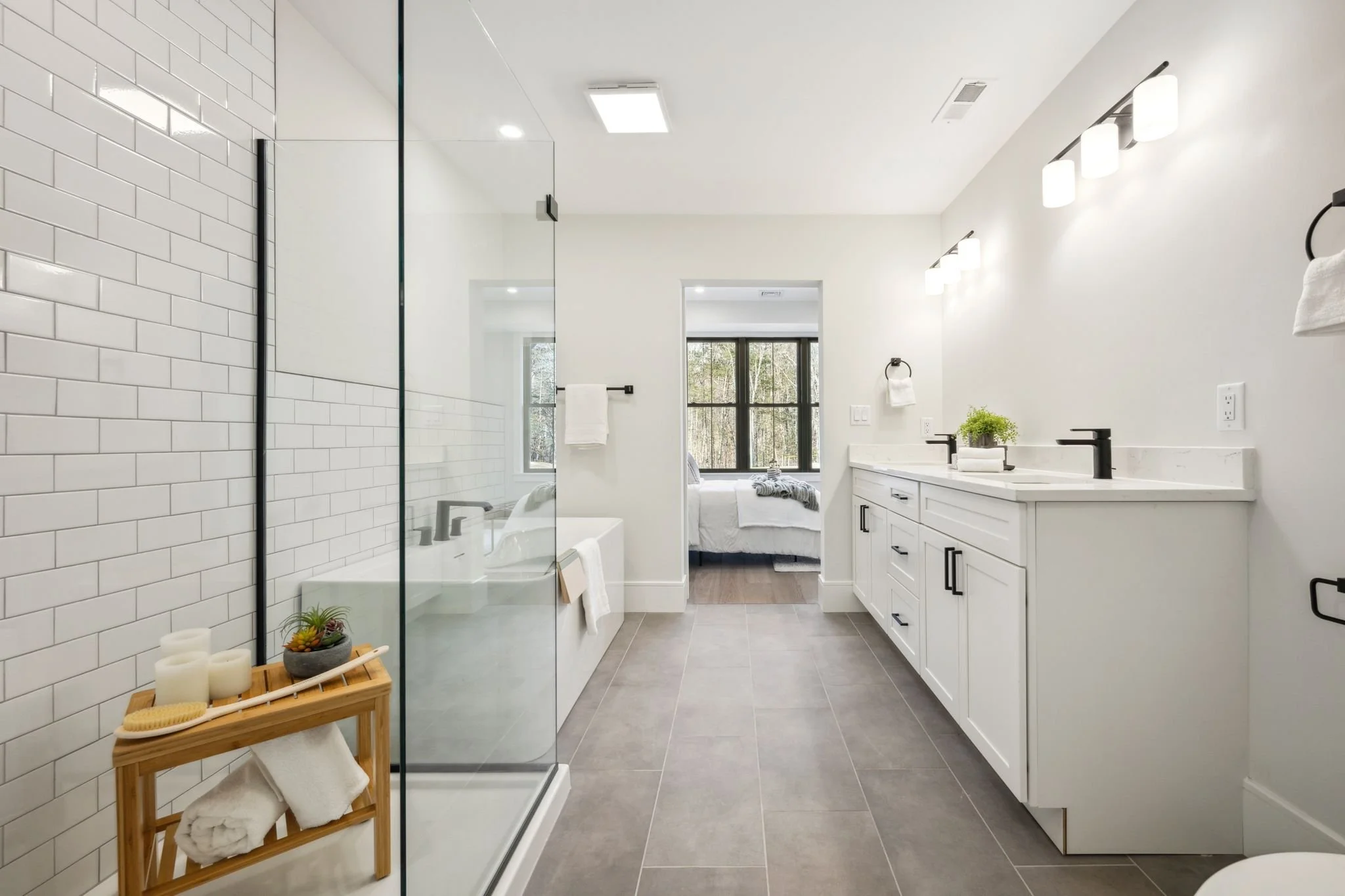 Modern bathroom with walk-in shower, white vanity with two sinks, and a view into a bedroom with a bed near a large window.