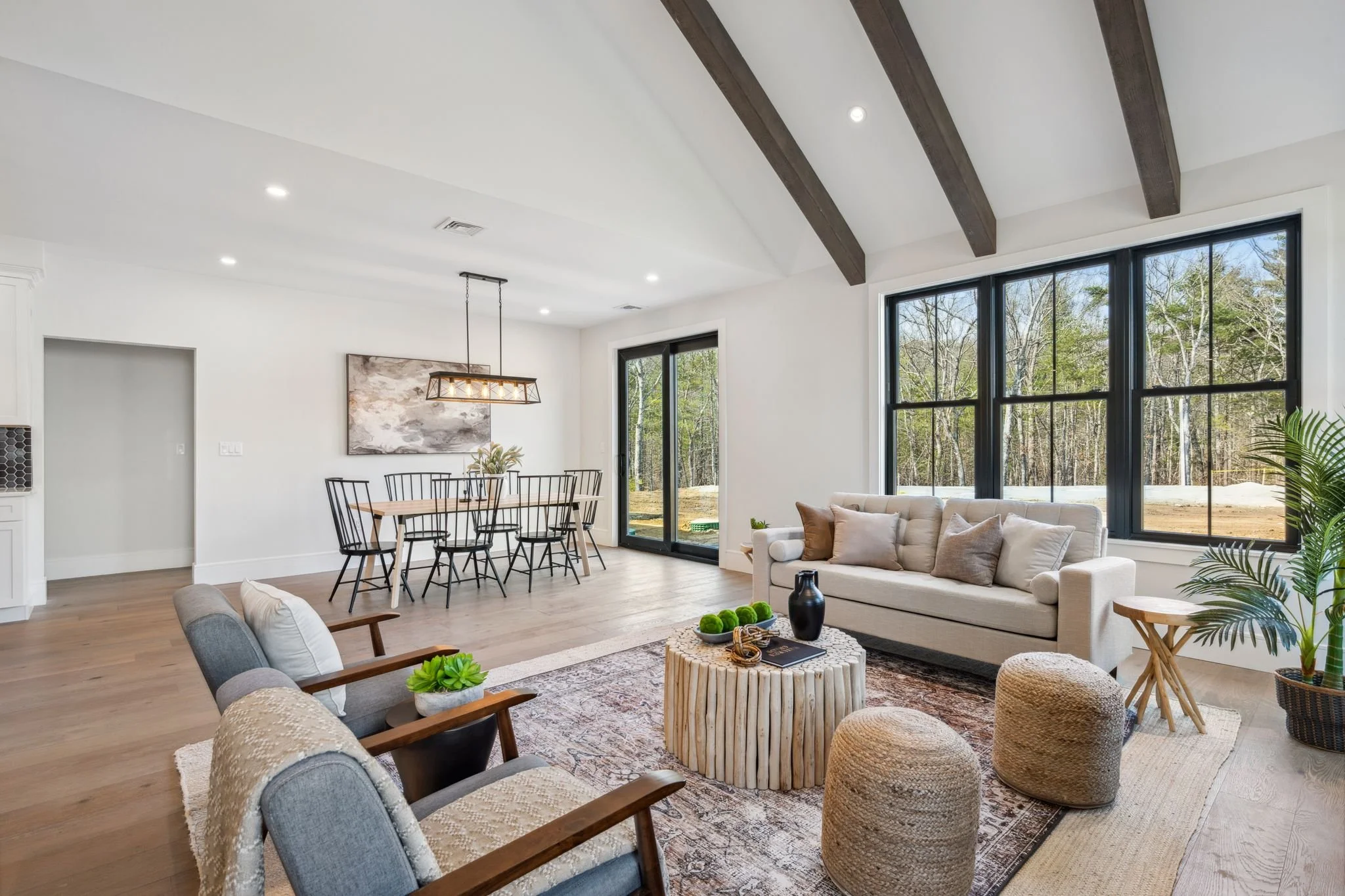 Living room with large windows, cream sofa, armchairs, wooden coffee table, and a dining area with a black modern chandelier, white walls, and wood beams on the ceiling.