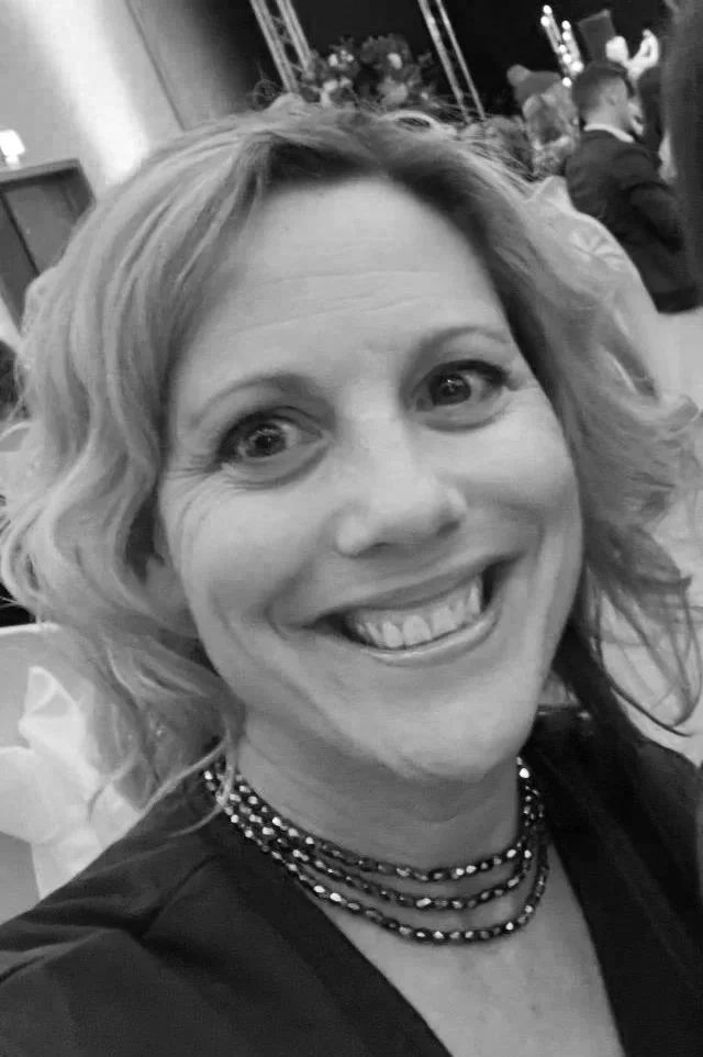 A woman smiling at a formal event, wearing a necklace, with an indoor background featuring other attendees and decorations.