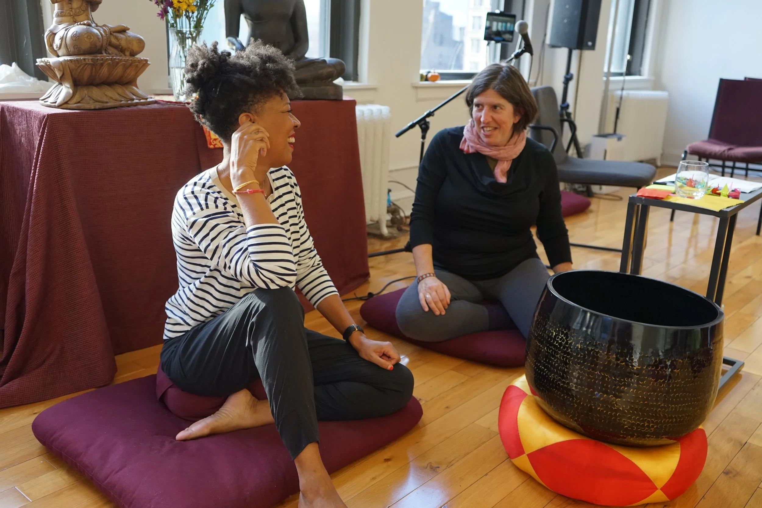 Two people sitting on floor cushions, engaging in conversation in a room with wooden floors and a window. One person is wearing a striped shirt and black pants, the other is wearing a black top and gray pants. There is a large black singing bowl on a