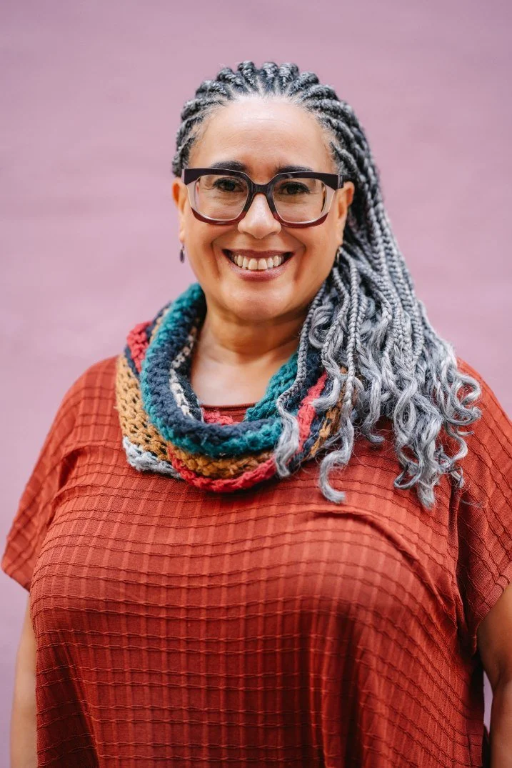 A smiling person with gray dreadlocks, wearing glasses, a multicolored knit scarf, and a red textured shirt, standing against a purple background.
