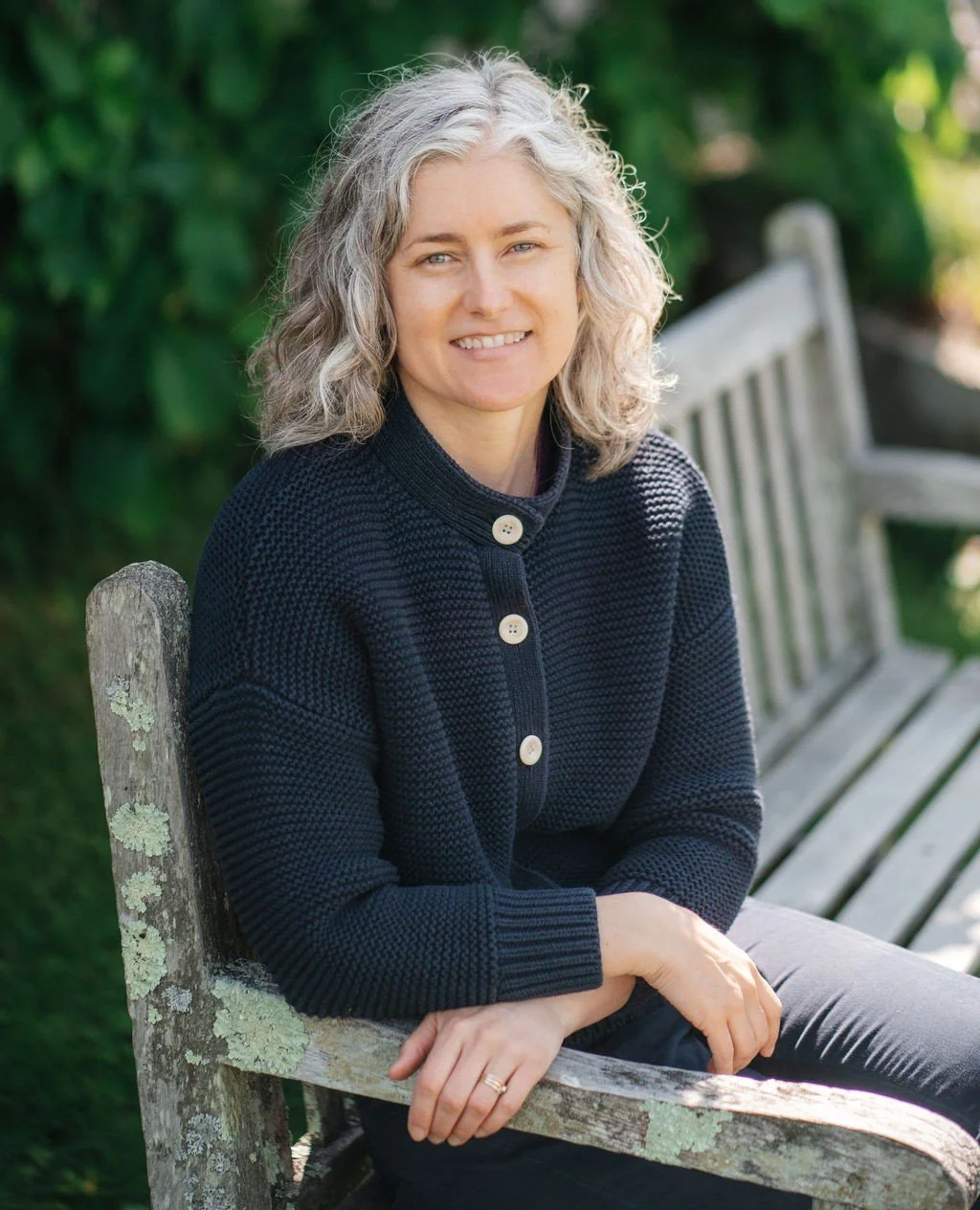 A person with curly gray hair sitting on a weathered wooden bench outdoors, smiling, with green foliage in the background.