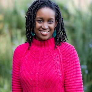 A person with dark hair wearing a bright pink sweater, smiling outdoors with green foliage in the background.