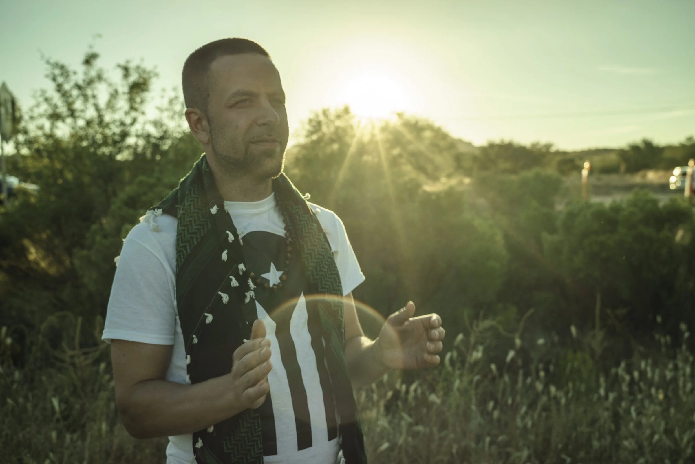 A person standing outdoors in the sunset, wearing a white t-shirt with black and blue graphic, with a scarf and beads around his neck, surrounded by greenery.