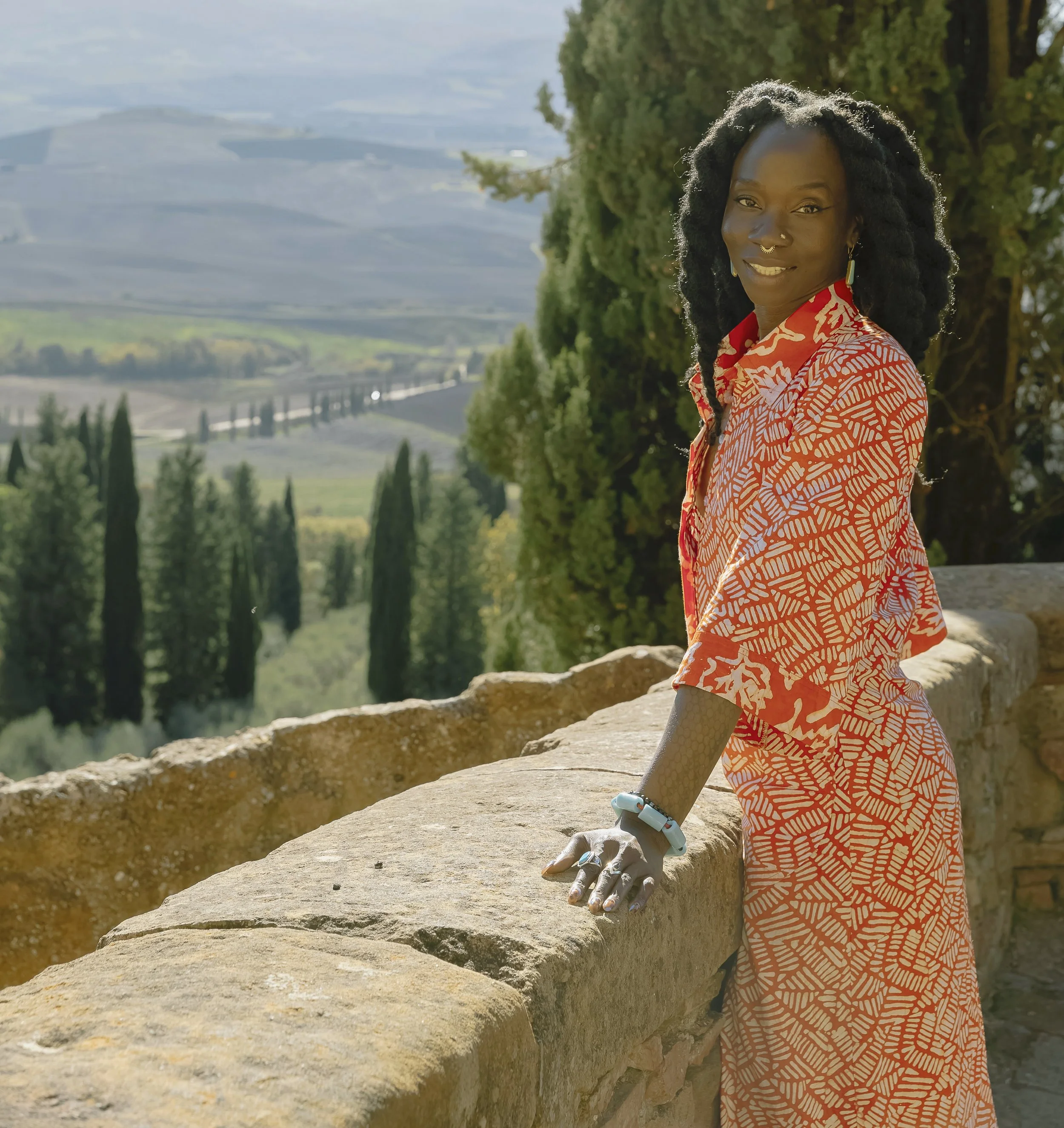 A person with curly hair wearing a red patterned dress, smiling, leaning on a stone wall outdoors with a lush green landscape and hills in the background.