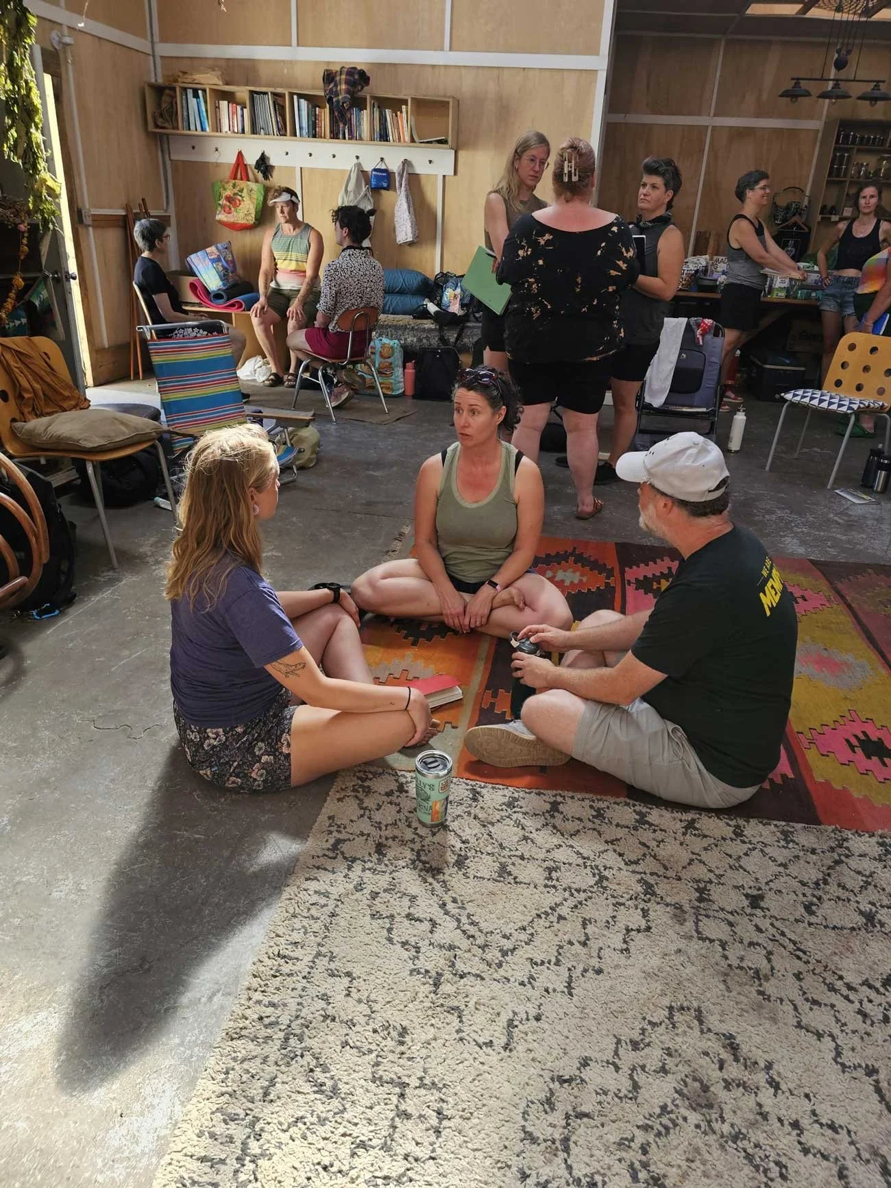 Group of people sitting on the floor in a circle in a room with bookshelves and wooden walls, engaged in conversation.