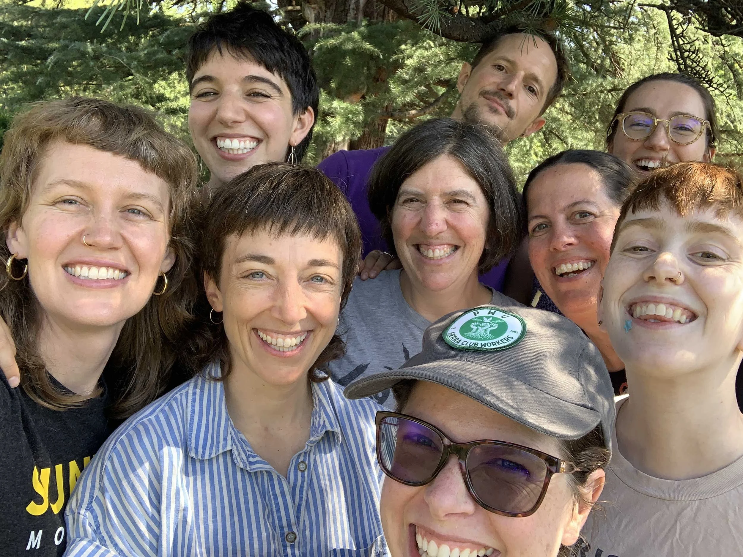 Group of 10 people smiling and gathered closely for a selfie outdoors, with trees in the background.