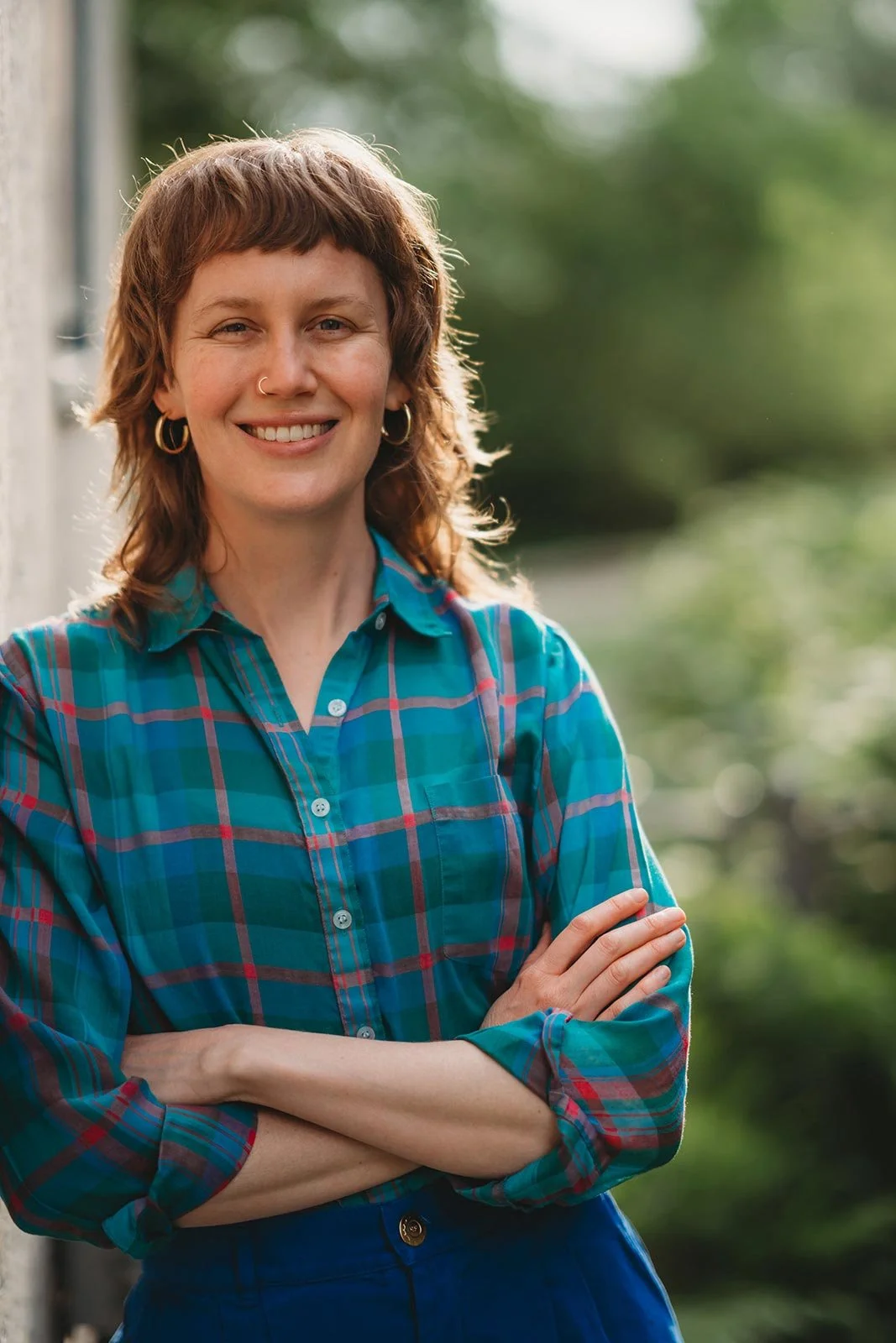 Smiling person holding purple flowers in a green outdoor space with trees and a house in the background.