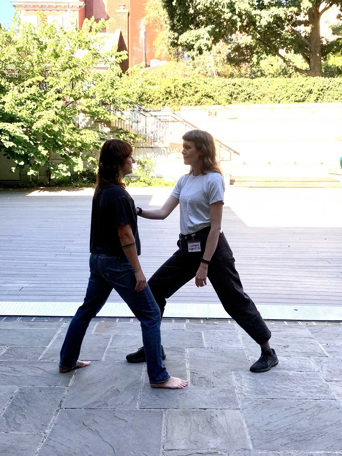 Two people practicing a martial arts demonstration outdoors, with one in a white t-shirt and black pants and the other in a black t-shirt and jeans, in front of a wooden deck and greenery.