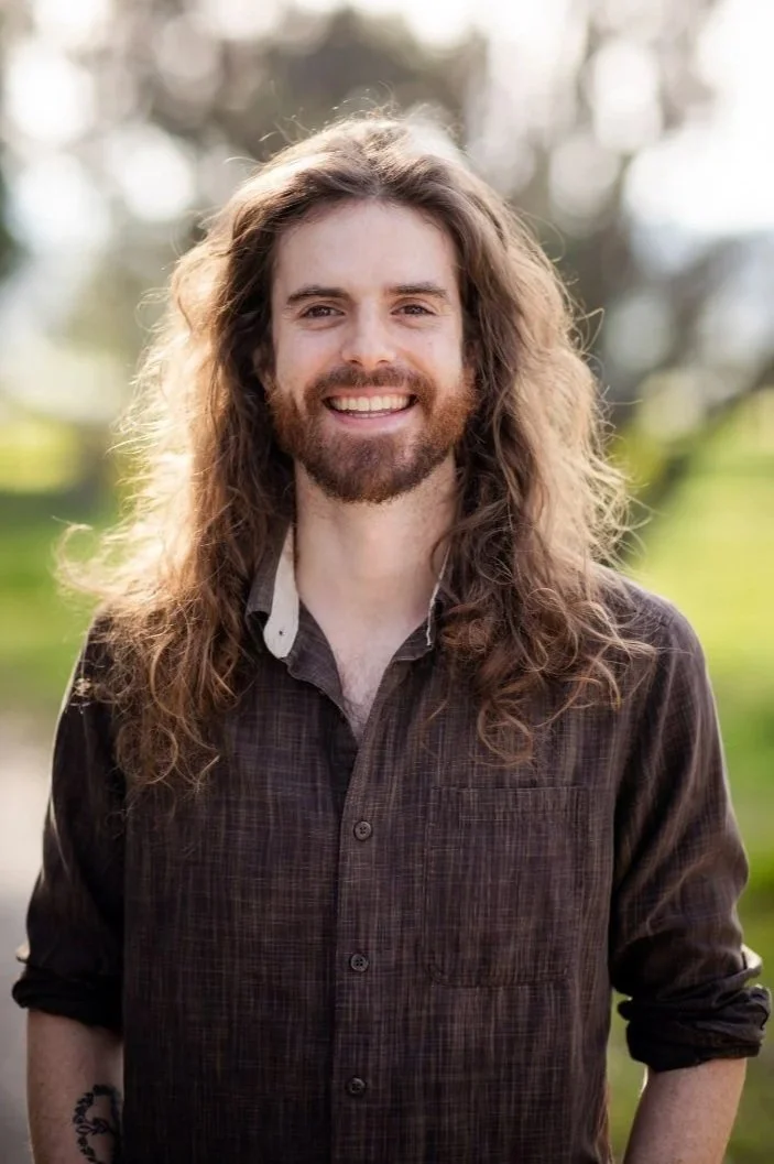 A person with long, curly brown hair and a beard is smiling outdoors. He's wearing a dark plaid shirt and the background is blurred greenery.