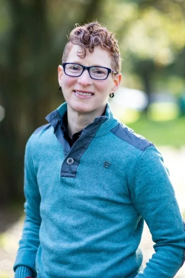 A person with short curly hair and glasses smiling outdoors, wearing a blue fleece jacket, with trees and sunlight in the background.