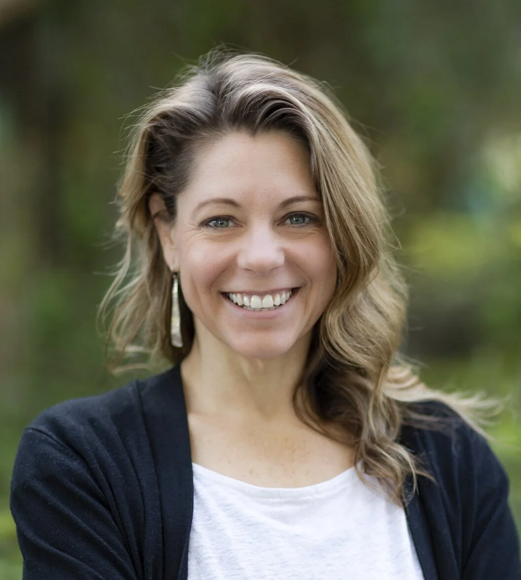 A person with curly brown hair smiling, wearing a white shirt and dark cardigan outdoors.