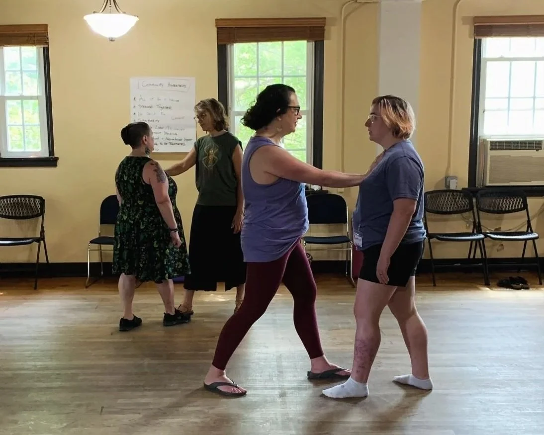Two pairs of people are practicing a dance or partner activity in a room with wooden floors and yellow walls, with chairs against the wall and windows.