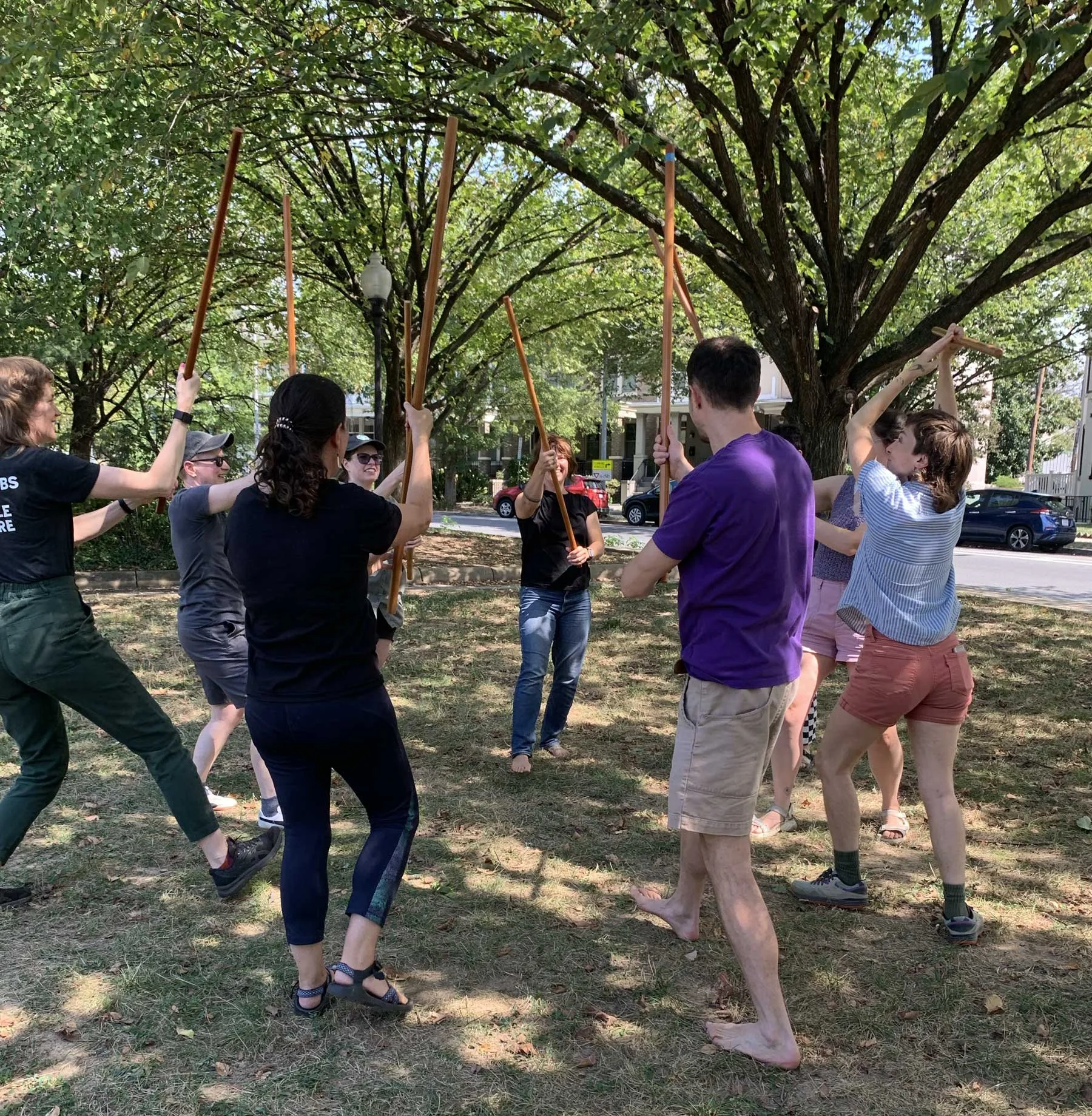 Group of people practicing jo kata in a park under a large tree.