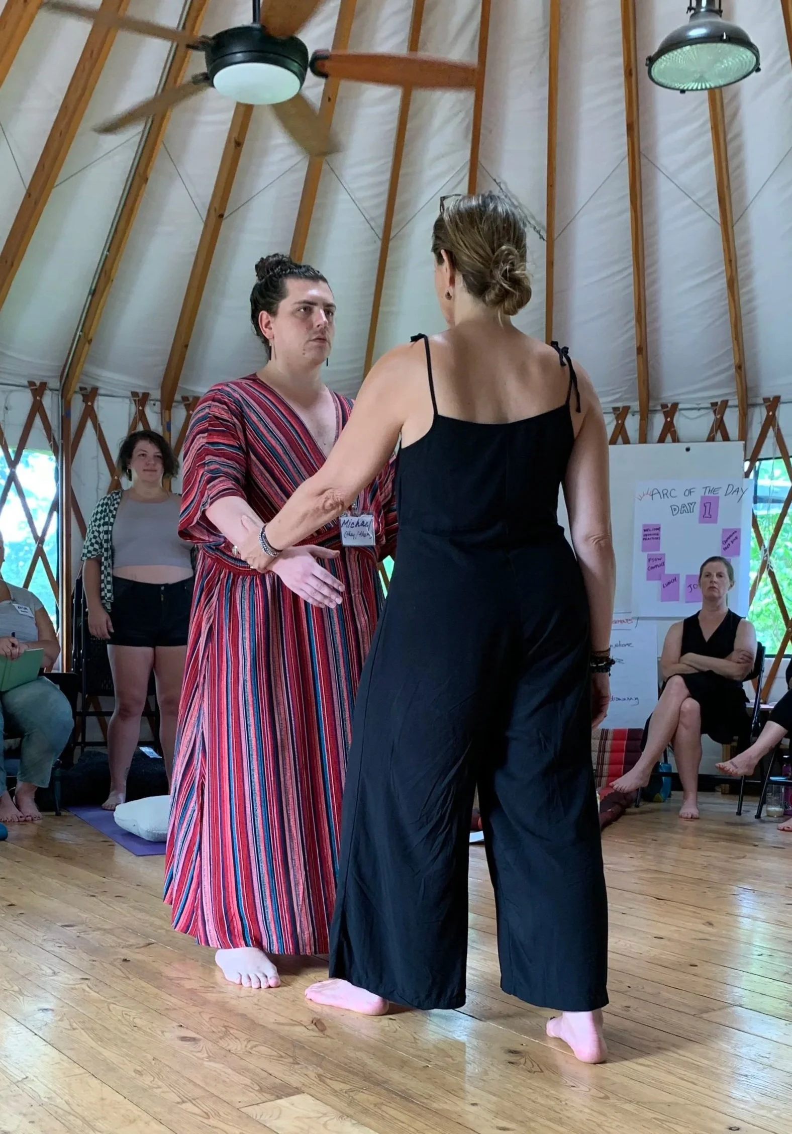 Two people are standing close together, holding hands and looking at each other. One person is wearing a long, striped dress, and the other is in a black jumpsuit. Several people are sitting and standing around in a wooden, tent-like room.