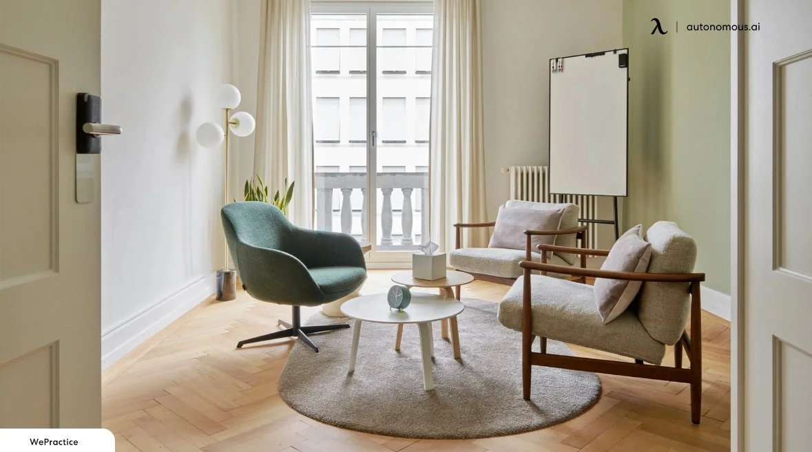 A modern living room with two beige armchairs, a green accent chair, and a small round coffee table on a circular rug, with large windows and cream curtains in the background.