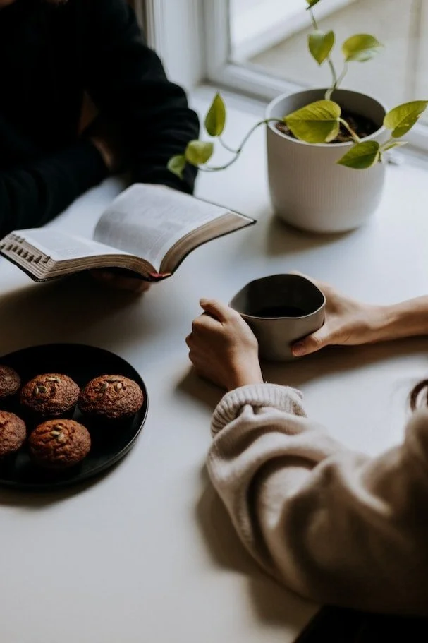 Two people sitting at a white table with a potted plant, a plate of cookies, an open book, and a cup of coffee or tea.