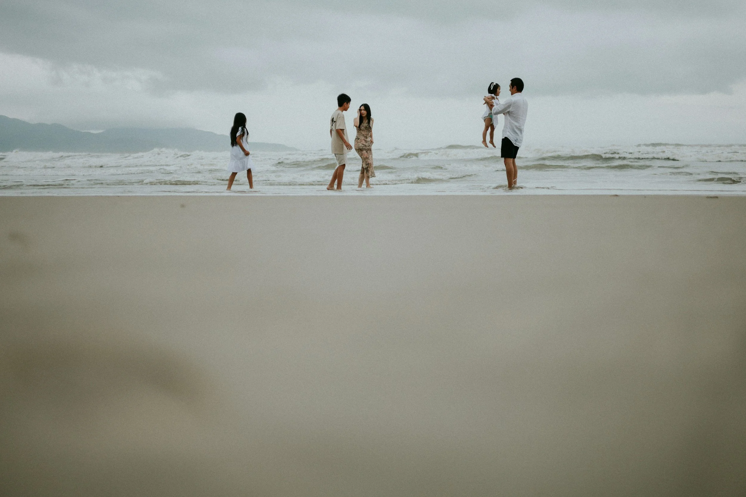 A family enjoying time at the beach with children playing and adults standing in the surf under gray clouds.