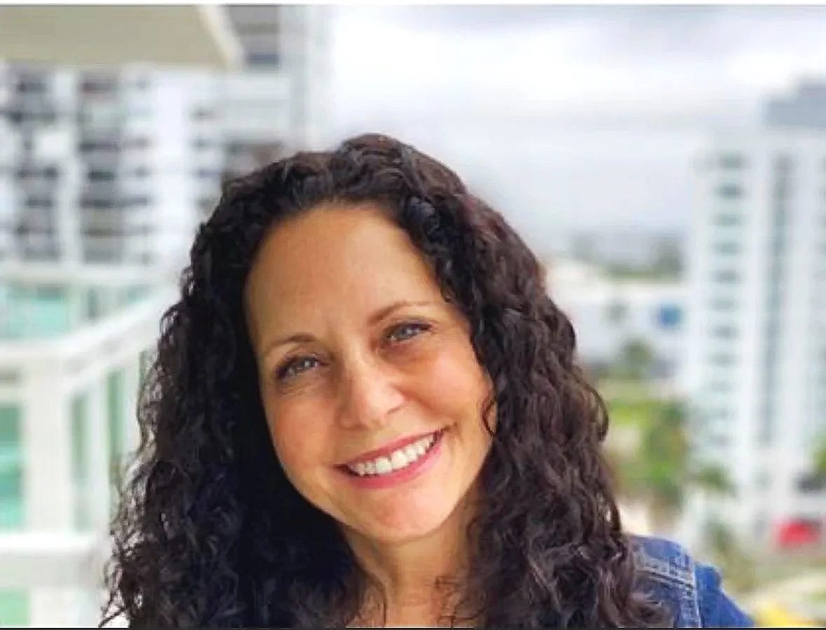 Close-up of a woman with curly dark hair smiling in front of a cityscape with tall buildings.