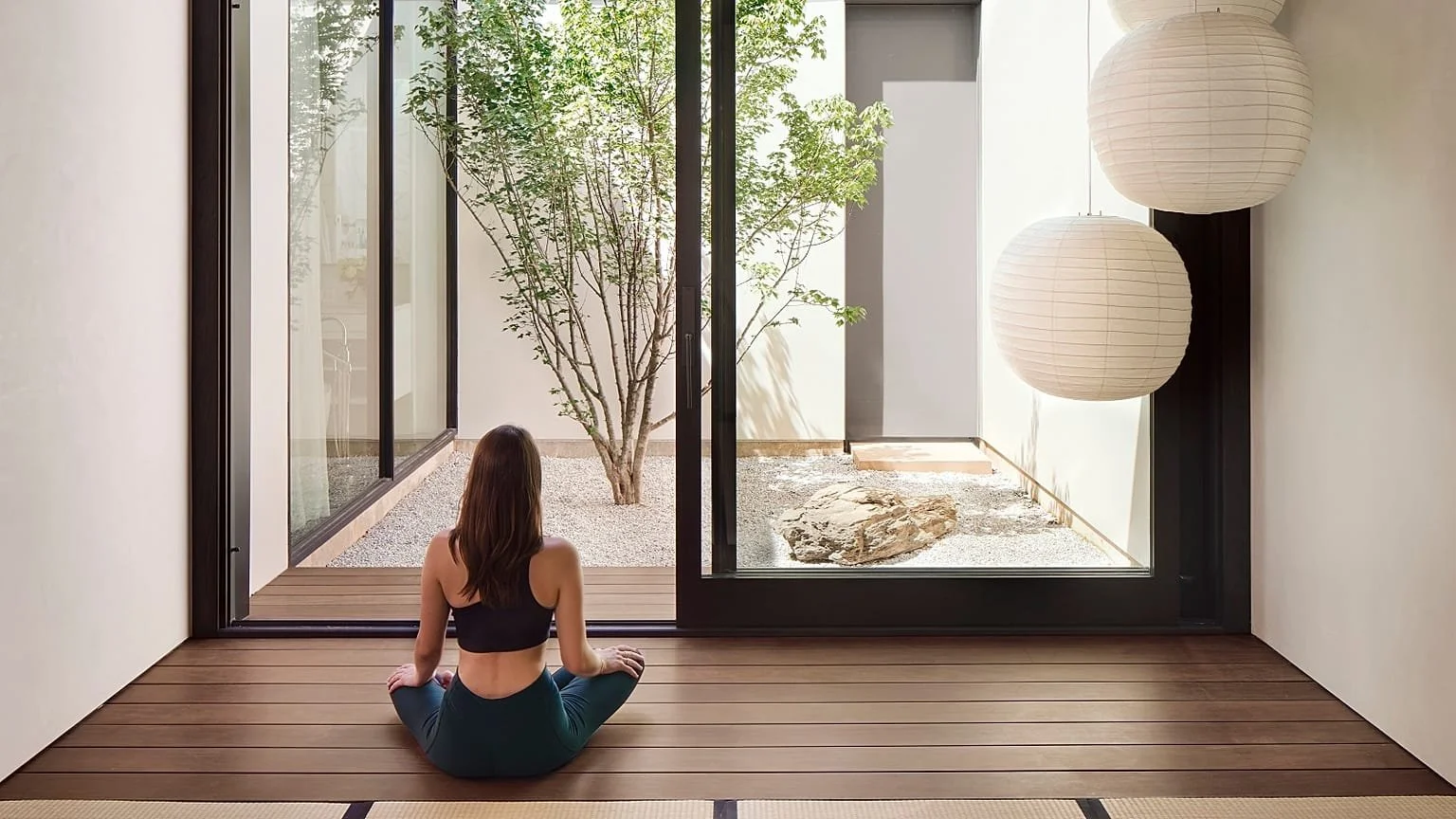 A woman practicing yoga in a minimalist room with large windows, a small garden with a tree, and paper lanterns.